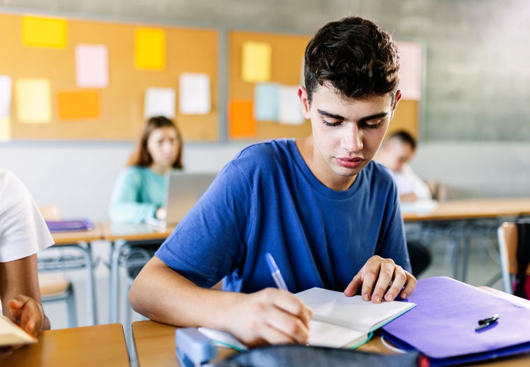 Jovem estudante do ensino médio escrevendo no caderno em sala de aula - Adolescente sentado na mesa fazendo exercício em sala de aula