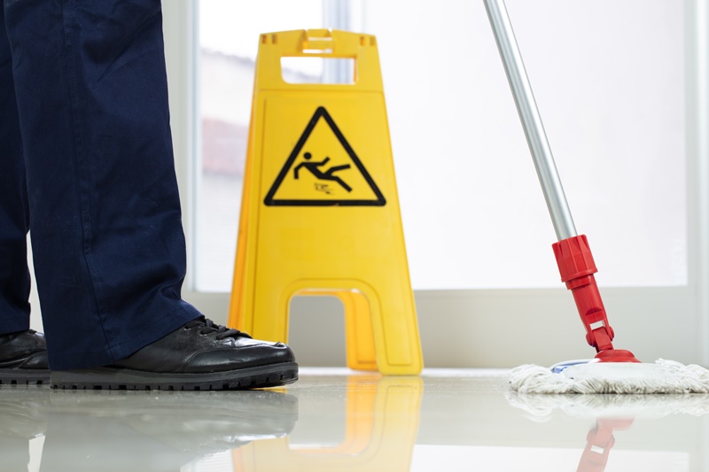 Closeup de baixo ângulo de uma pessoa limpando o chão com um esfregão perto de uma placa amarela de advertência sobre piso molhado, representando o tratamento de piso em supermercados