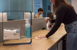 selfcleaning da rl higiene que trás limpeza para empresas. na foto uma mulher limpa uma mesa, estação de trabalho em escritório enquanto um homem faz seu serviço no fundo