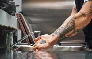 homem praticando o ato de lavar as mãos, lavando-as em uma pia de restaurante, no braço dele há diversas tatuagens