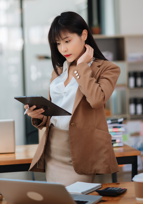 Empresária asiática de terno beje e camisa branca, cabelos pretos analisando o manual de etica da rl em tablet e laptop no escritorio